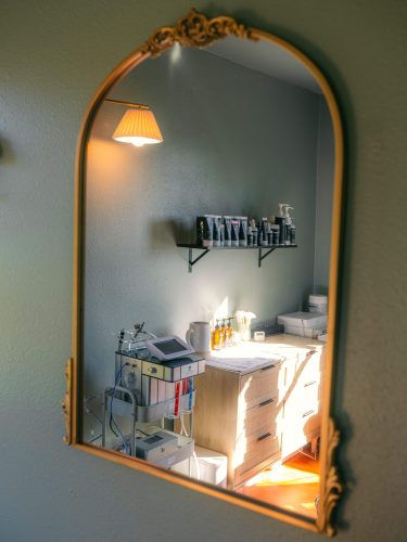 Gold arched mirror reflects the sunny facial treatment room and skincare product display.